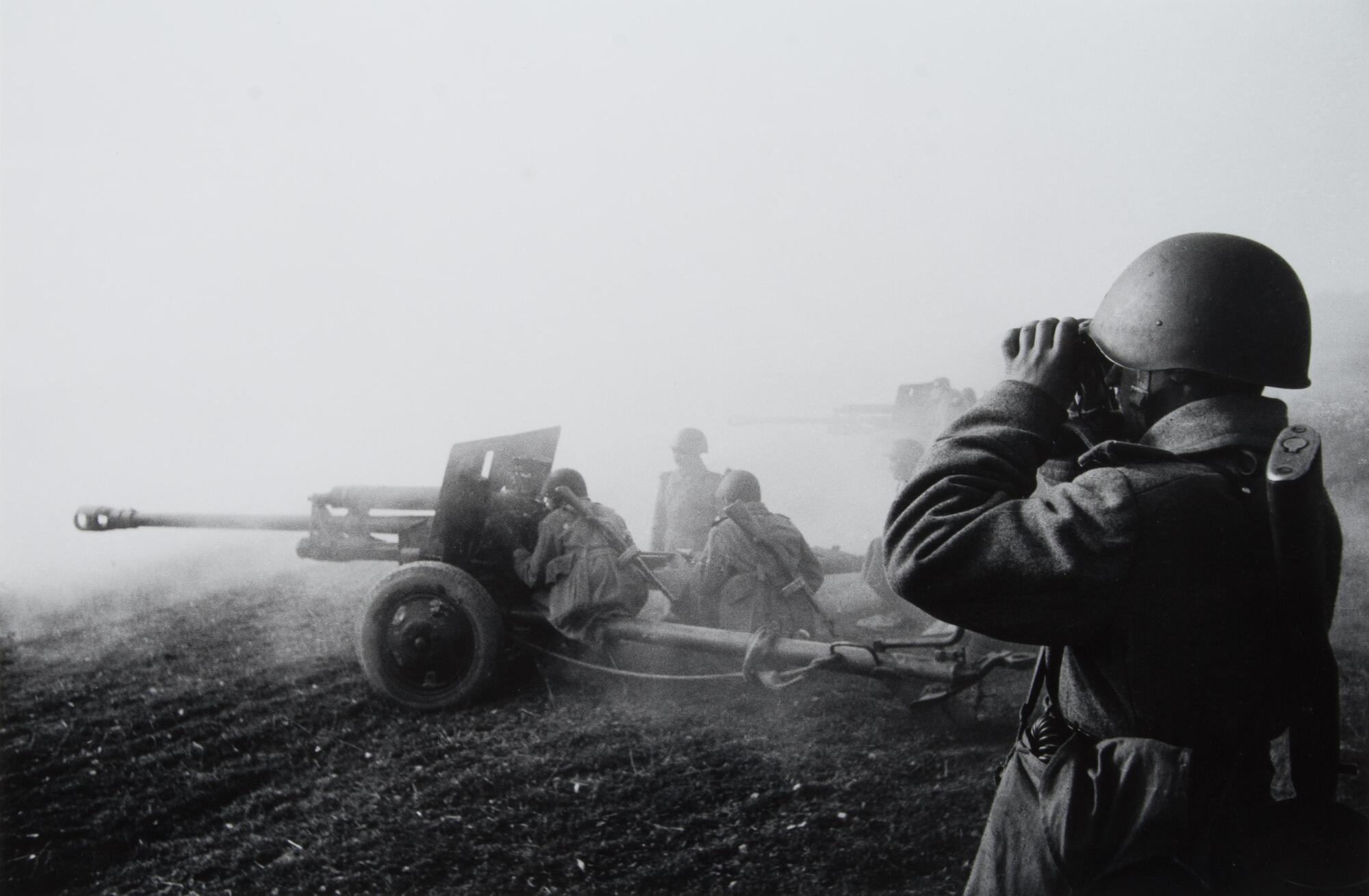 In the lower-right foreground, a soldier with a round helmet peers through binoculars. In the middle ground, other soldiers man an anti-tank gun while fog and/or smoke fills the upper register of the photograph.