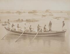 This photograph shows a view of nine men, three of them oarsmen, on a small boat currently paused in shallow water.