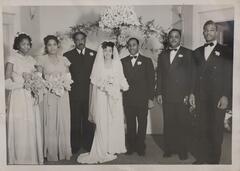 A bride and groom stand under a flower-covered wedding altar at the center of the image, flanked by two formally-dressed men on the right and a man and two women on the left. The bride wears a white dress with a veil and holds a bouquet of flowers. The women on the left are also wearing dresses and holding bouquets.