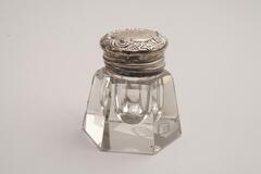 This is a cut crystal inkwell with a sterling silver lid. The body is a hexagon shape with cut shoulders. The top has a rounded shape and floral designs.<br />