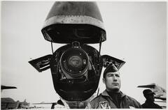 A uniformed man seen from the shoulders up stands at the nose of a jet that has been opened and raised to reveal its inner workings.
