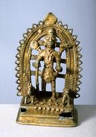 Vishnu stands with his legs apart holding his four attributes in his hands.  Reading in clockwise direction from his right front hand he holds:  his club, discus, conch and lotus, here a rather flat object cupped in his palm.  His back two arms are extremely short. The figure is encircled with a decorated arch with a line of beads and triangular shaped openings around them.  A stylized sun and moon are to either side of Vishnu’s head.  He wears a variety of simple, lumpy jewelry at his feet are a horse to his right and a bull or cow to his left and between them are three rings lying flat on the base.  At the front of the base are seven stylized horses, identifying this as a combination figure: Vishnu and the sun god Surya, whose chariot is pulled by seven horses.<br />