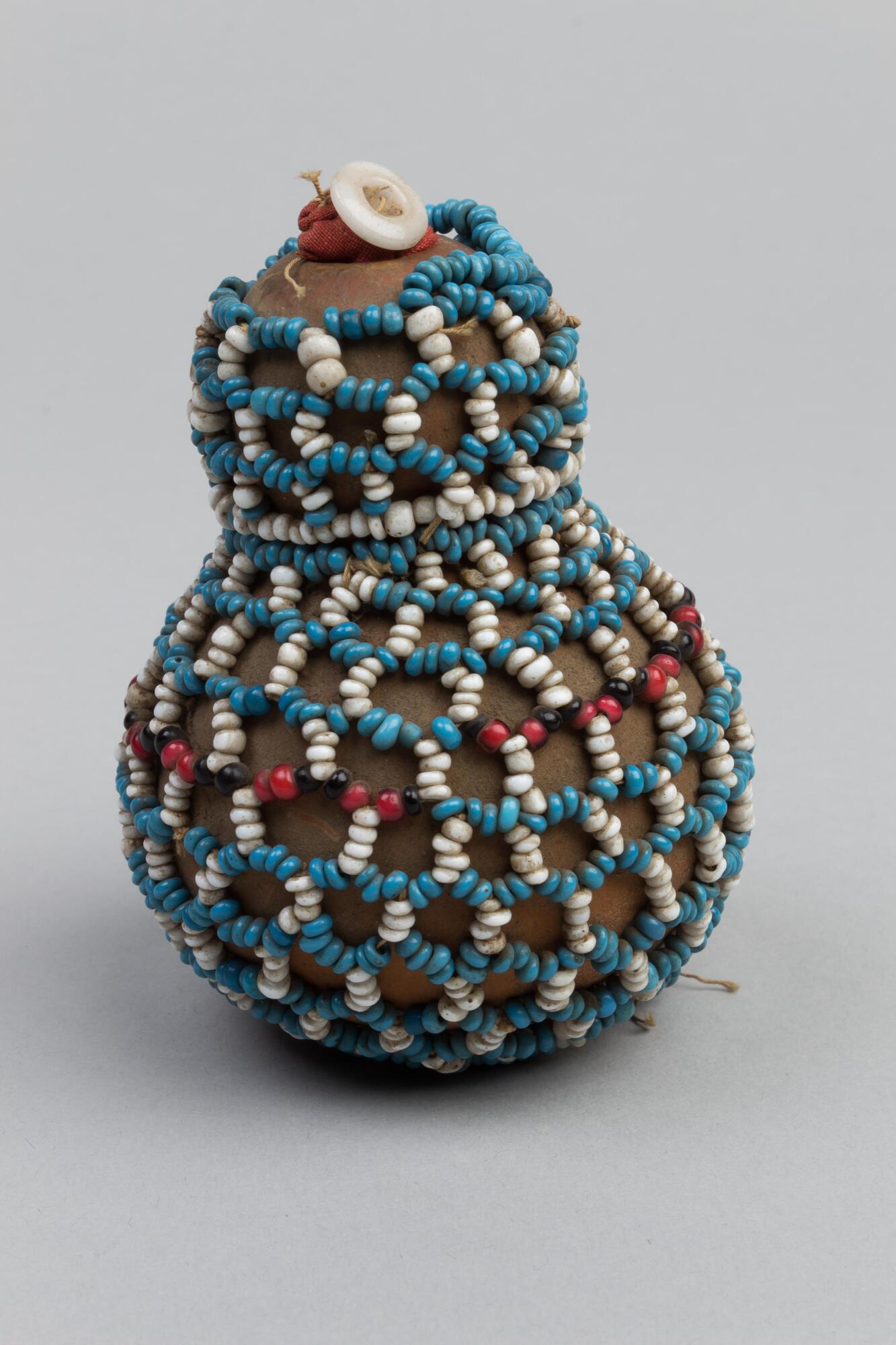 Gourd with beaded open weave cover in blue and white with some small short black and red detail. Gourd is filled with red fabric with buttons at top. Short blue beaded handle at top, white tag attached.