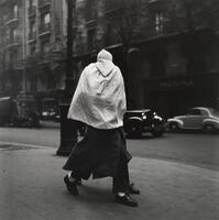 Two people walking along a street, cars in road. One person is carrying a baby wrapped in a blanket and hat.