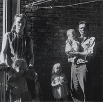 A portrait of a family standing outside in an alleyway. From left to right, a woman stands with her hands on her young daughter's shoulders, while a young boy, barely visible, peeks from behind her. Another young girl stands in the center next to her father, who holds an infant on his hip.
