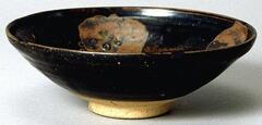 A shallow, wide bowl with a wide bottom on a tall straight footring. It is covered in a dark black-brown glaze, with four russet overglaze iron oxide splashes to the interior. There are chips to the rim.