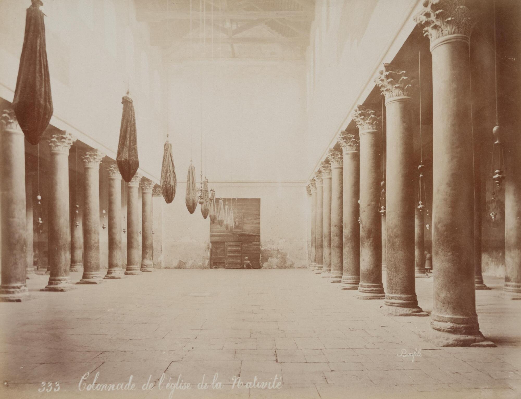 Interior view of a mostly deserted basilica, lined on both sides by rows of columns. A figure sits on the ground with his back against a wall made of wooden slats. 