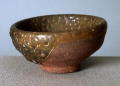 A shallow bowl on a tall straight foot ring, covered in a thick brown-ochre glaze. The too-thick misfired glaze crawling and blistering away from the underlying clay body.