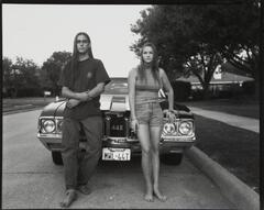 A boy and girl (both with long hair), sitting on the hood of a car. Both are barefoot and the car has a Texas license plate.