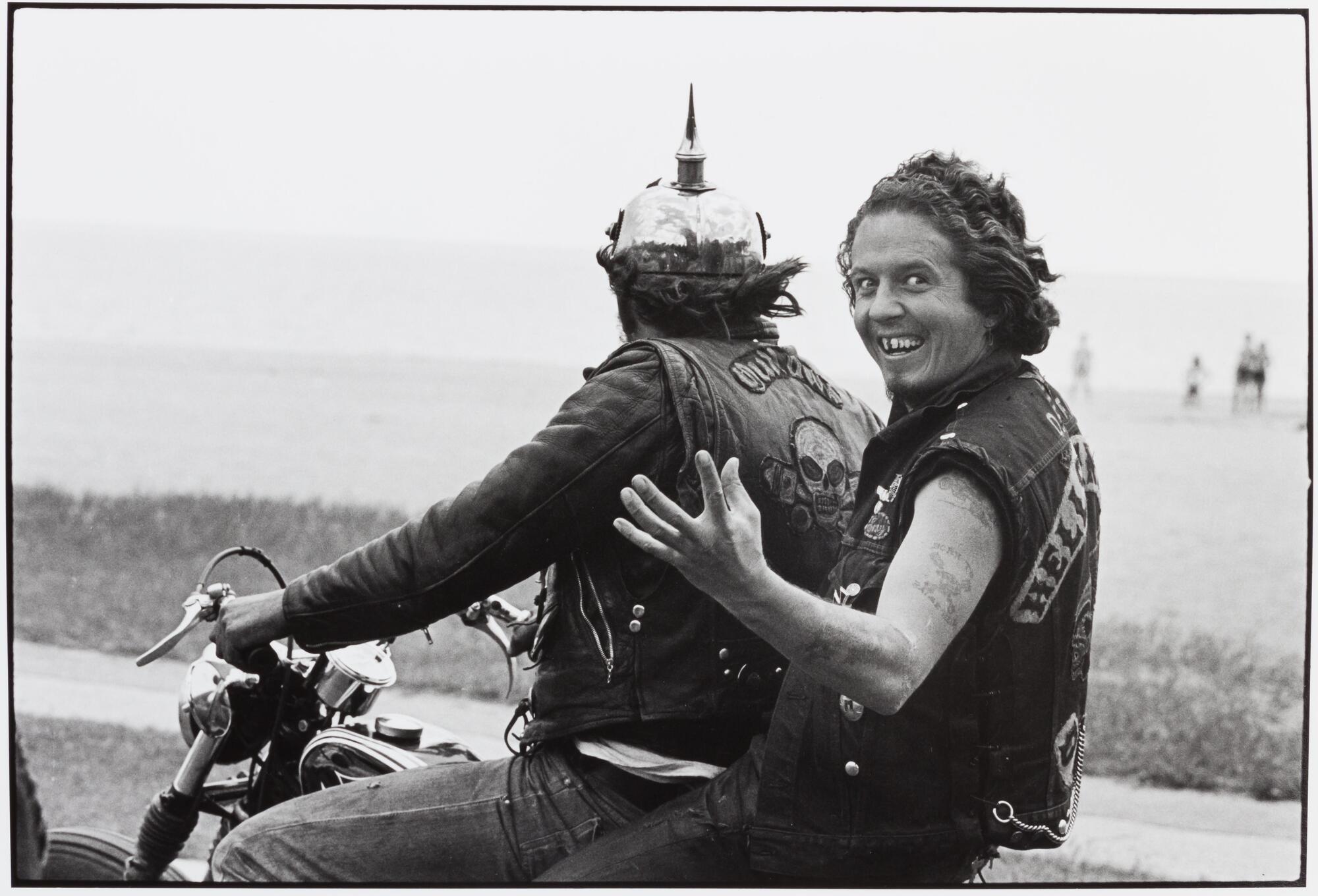 A photograph of two men riding on a motorcycle. The man in the back seat looks back at the camera, holding out his hand and smiling. The other man wears a helmet with a large spike and looks toward the beach scene that unfolds before him.