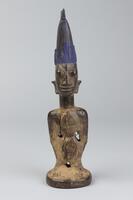 Standing male figure on a round base with its hands at the sides and a triangular pendant on the front and back of the chest. The pendant is also on the sides of the hair, which is tall, topped by two small triangles, and coated in blue pigment. 