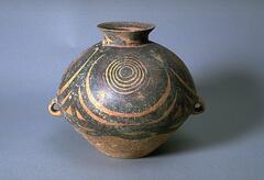 A light reddish-buff earthenware <em>guan </em>罐 jar with a wide globular upper body and conical lower body on a flat base, and a short narrow everted neck with direct rim. There are two diametrically opposed lug handles at the waist. The upper half of the body is painted with black and red pigments to depict four whirls of concentric circles, each containing one red circle, confined between solid band borders, with a garland border below. Around the neck is a thick chevron pattern, with a red band and black ladder pattern band to interior rim.