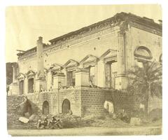 This photograph depicts a view of a devastated palatial building. Before its shattered portico is a pile of rubble and fragments of columns. Four men in turbans are seated on the ground before the building. 