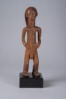 Carved wood figure of a human. The figure's torso, neck, and head are cyclindrical and elongated on the vertical axis. The figure's arms rest by its umbilicus, which protrudes slightly. Details of the hair, face, hands, genitals, and feet are carved in. The figure is incised with symmetrical, zigzag-ing lines.