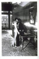 This photograph depicts two models standing in an elegant interior. They are embracing and kissing, posed to appear as mannequins. 