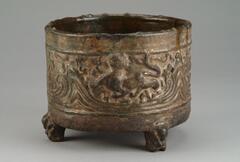 Red earthenware cylindrical vessel on three zoomorphic feet, molded in low relief with hills and running animals, covered in a lead green glaze with iridescence and calcification.