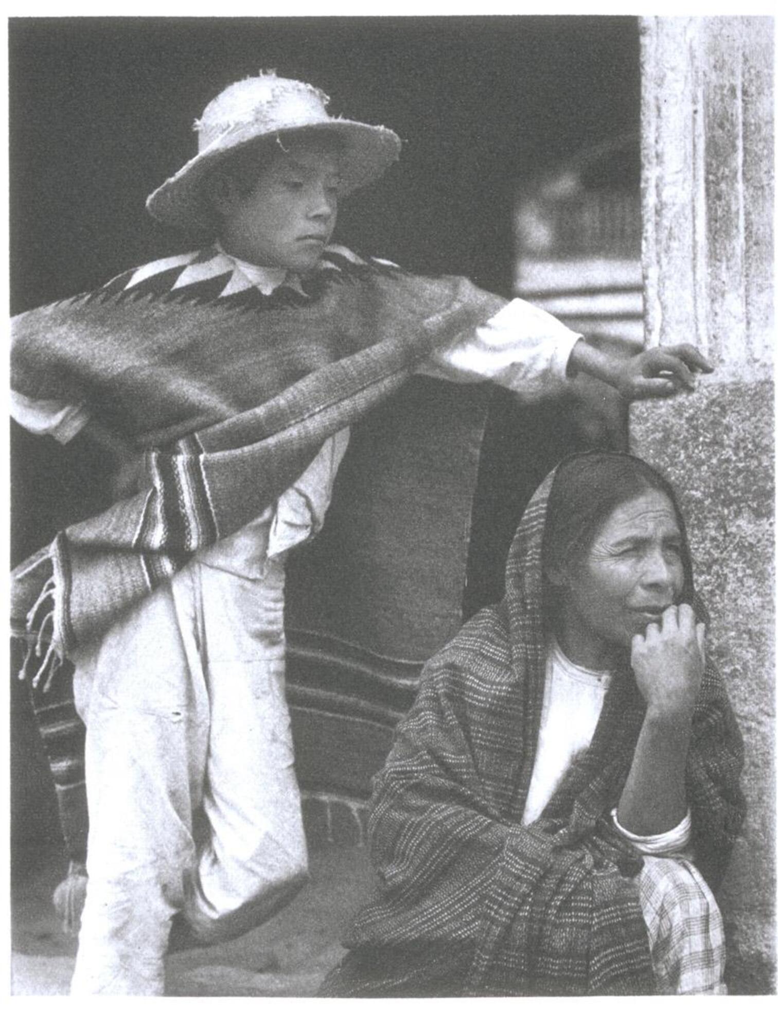 Portrait of a young boy and woman. The boy stands with his right arm akimbo and his left arm resting on the corner of a building. The woman crouches next to the building, with her head just below the boy's arm.