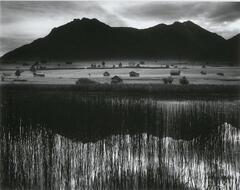In the foreground, a body of water framed by tall reeds reflects the mountain range behind it. Beyond the lake, small buildings dot the landscape as mountains and sky rise above them in the background.