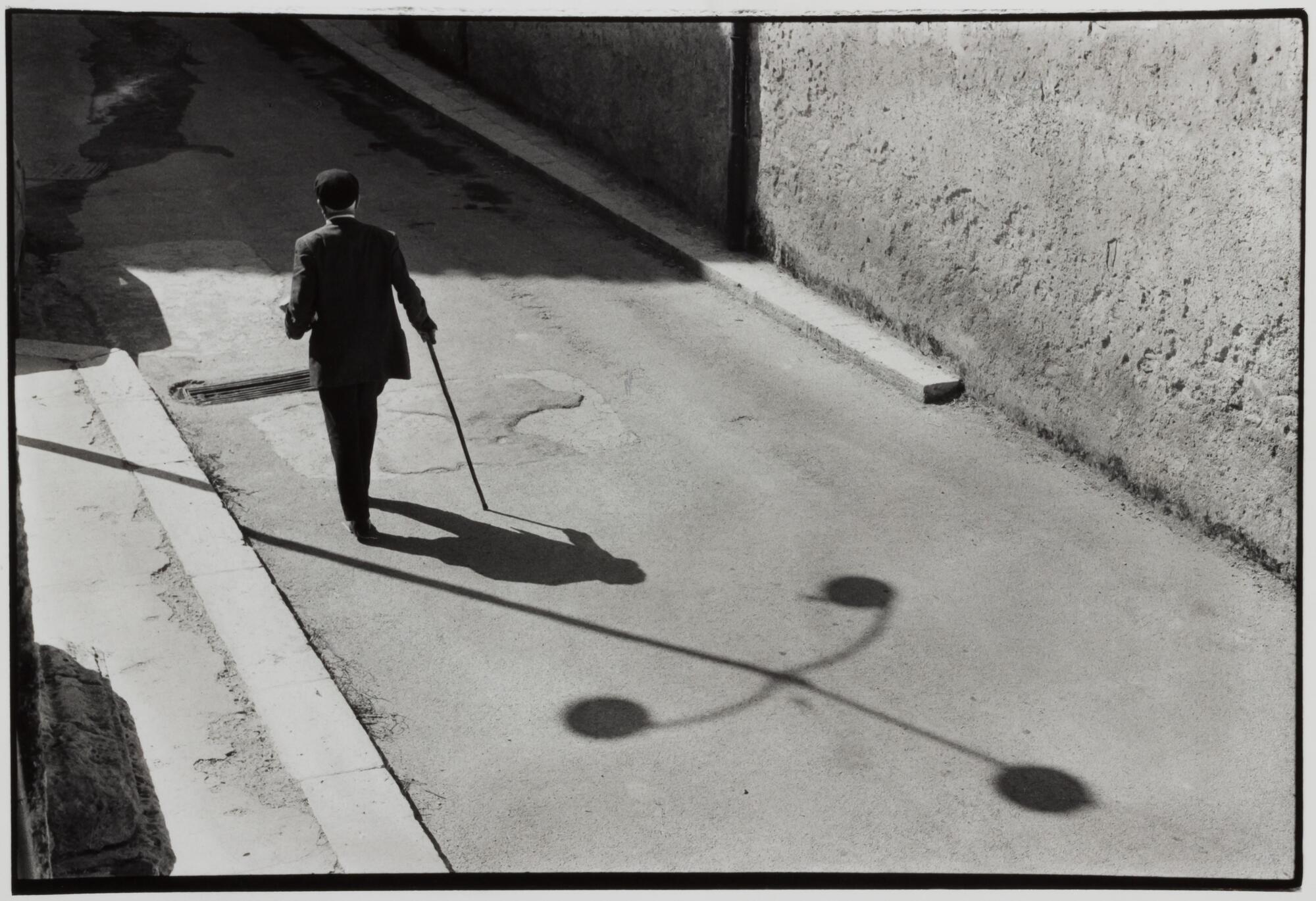 A man walking down the street using a cane. There are shadows reflected on the sidewalk.