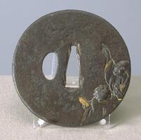 Circular tsuba, made of iron. It has two holes in the middle. Two figures, Kanzan and Jittoku, are carved on the lower right corner. Kanzan, who holds a scroll on his hand, and Jittoku, who holds a bloom stick and pointing to the sky, are looking upward. The two figures are carved slightly higher than the surface. On the back, there is the moon partially obscured by clouds. Gold and silver alloy inlays are applied to the moon and the clouds. Gold is also inlayed in their eyes, parts of the garments, and Kanzan's scroll. Shakudô (copper-gold alloy) is inlayed in Jittoku's bloom and his jacket collars.