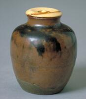 Small, brown, thinly potted container with ivory lid. It has a short neck and round shoulder; The dark glaze is randomly applied from shoulder to the middle of the body. The part at the bottom is unglazed. The bottom has no foot and unglazed. The lid is concaved from the rim toward the center knob.