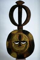 Circular mask with a round face in the center and a circular superstructrure. The eyes are cresent-shaped and the forehead and outer edge of the face are raised. The edge of the mask is decorated with alternating semi-circles in yellow and black pigment. The face of the mask is yellow with a dark marquise shaped mark in the center of the forehead. Above the central face is an open circle intersected by a vertical bar decorated with a spiral design. At the very top of the circle is a diamond-shaped projection. 