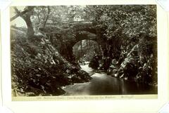 In this photograph, an arched bridge is depicted surrounded by trees with a rock-lined river below. 
