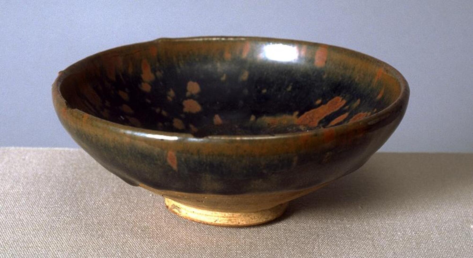 A wide, shallow, bowl on a straight foot ring, covered in a thickly applied, dark, iron-rich black glaze with lighter russet-brown patridge feather markings (鷓鴣斑 <em>zhegu ban</em>) .  The thick glaze thins at the rim to a russet-brown color.