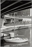 A man on a sailboat underneath a highway overpass. There is another man standing on the dock. There is a large office building in the background.