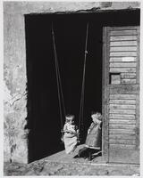 Photograph of a young girl standing next to a baby in a swing that hangs from the upper frame of a doorway.