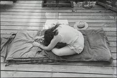 Girl leaning over while seated on top of a rolled-out sleeping bag. It is on a wooden deck and there is hat on the boards.