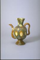 This is an earthenware globular ewer on a tall flaring footring. It has a tall flaring neck with a coil ear-shaped handle extending from shoulder to belly. An "S" shaped spout extends up from the lower belly, which is stamped with floral designs and covered in green, amber, and cream glazes. 