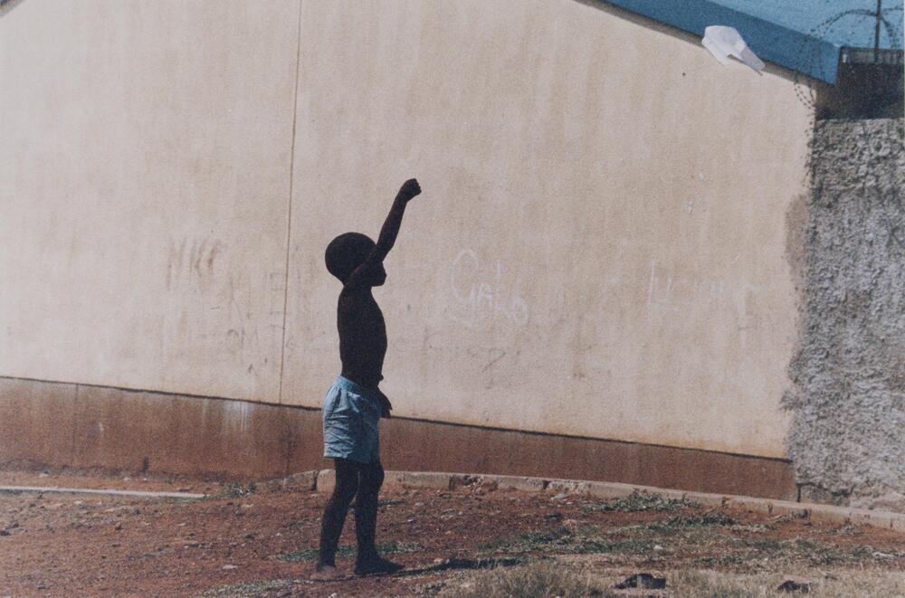 A young boy standing next to a long wall with grafitti with his left hand raised.