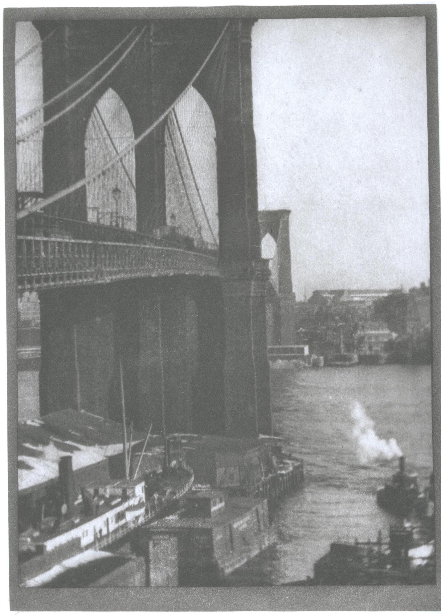 This photograph depicts an oblique view of the Brooklyn Bridge. The bridge dominates over half of the frame starting on the left and reaching into the background, while the right side represents the river and boats underneath the bridge.