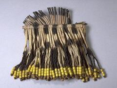 An apron with columns of woven fiber hanging from a wooden rod that have alternating patches of dark and light coloring. Attached to the bottom is a fiber fringe with alternating light and dark coloring with yellow beads. The top of the apron also has small stiffened fiber rods with alternating patches of light and dark coloring.