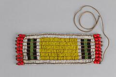 Beaded patch. Main section in small white beads with yellow rectangle in small beads and some green and blue stripes. Red beaded tassles on either end. Brown thread. Loose long thread coming off end.
