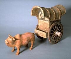 This is an earthenware model of an ox and covered cart. The ox stands on all four legs and is hump-backed with horns. He is painted in red pigment and stands in front of a two spoke-wheeled cart, which is closed and covered and painted in red, black, and white pigments. 