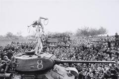 A man and woman perform circus tricks on top of a stopped tank while a crowd of soldiers watch. 