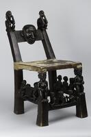 This wooden chair has two squatting figures decorating the finials; one is elderly, bearded, and scarified, while the other is fresh-faced and young. At the lower left rung, two men carry a slit drum; between the caryatid figures supporting the chair’s front legs appear three men; the central slat has incised diagonal patterns called fuliko. At the center, what is possibly a pointy-nosed European’s face replaces the more habitual chikungu masker.