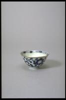 A petite cup with an abstract floral design in blue and white covering the outside of the cup and inside rim. The outside rim is gold and slightly everted. It has a large foot with blue stripes.