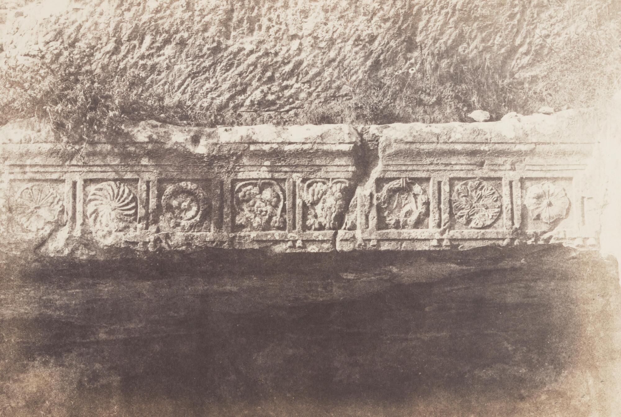 Photograph of a large stone carving occupies the center of the image. Above is rough-cut stone, indicating that the carving is part of a larger stone setting; below is a recess and darkness, indicating that the carving acts as a kind of decorative lintel above a cave entrance. The carving itself consists of eight square zones (or metopes) and within each is a decorative pattern of radial symmetry (circles) or vegetative imagery. The frieze is located above the entrance to the Apostles' Retreat, Valley of Hinnom in Jerusalem.