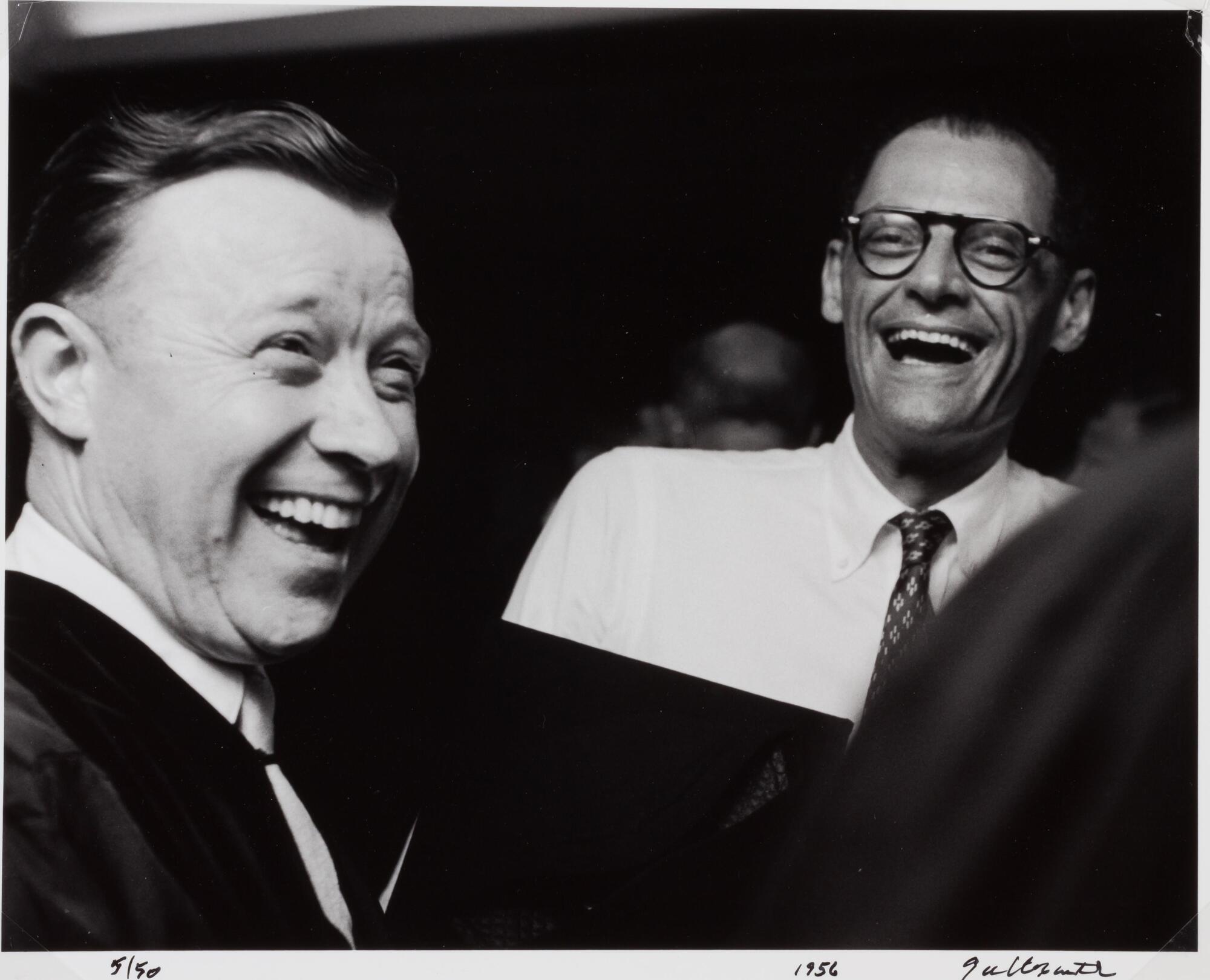 Two men standing next to eachother, both are laughing. One is wearing a suit jacket, the other is in a white shirt and tie with glasses.
