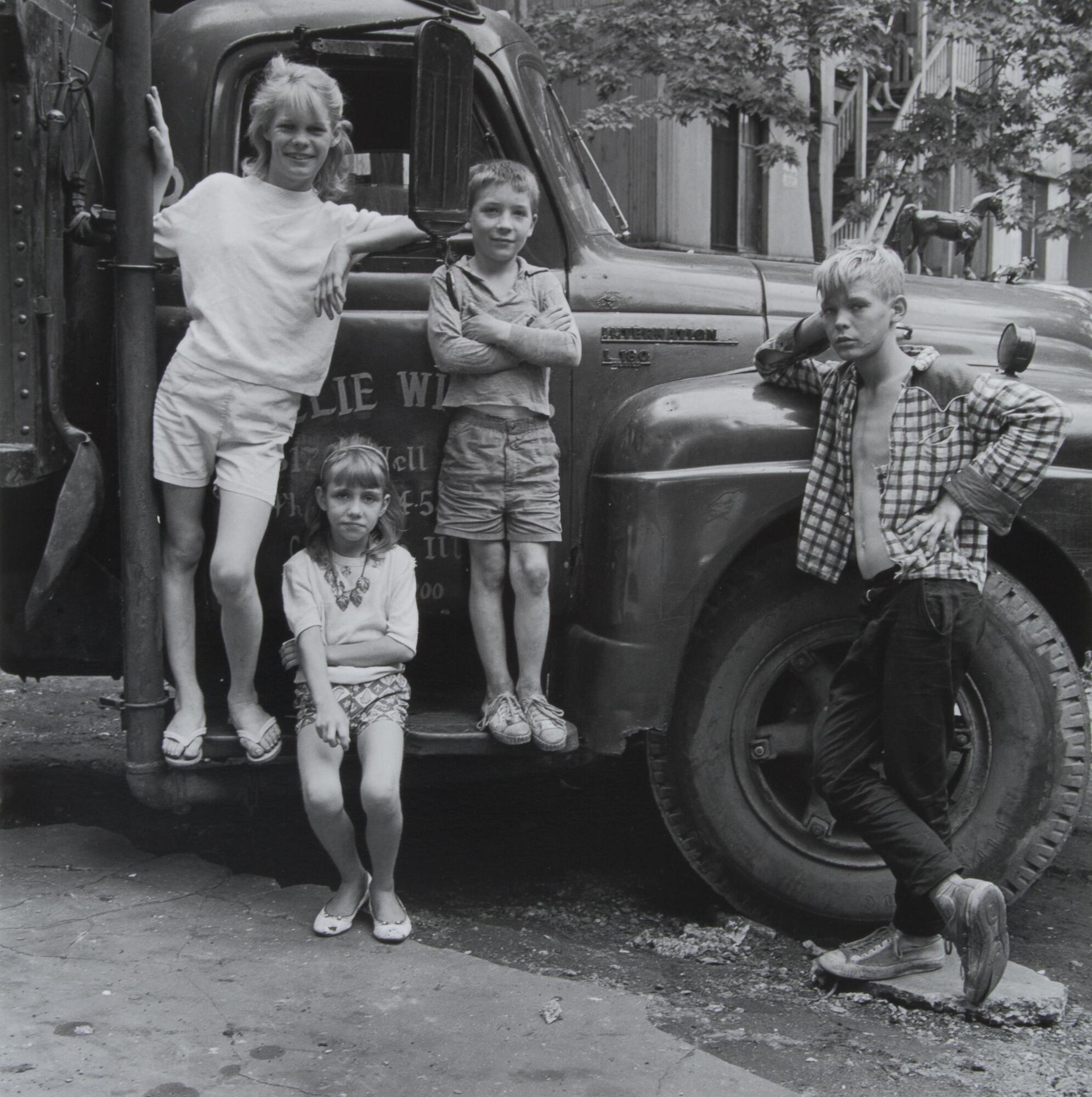 A photograph of four children leaning against a truck. On the left, three children are grouped together, two young girls, and a boy. On the right, another boy leans his arm against the hub of the wheel; his other arm is akimbo.