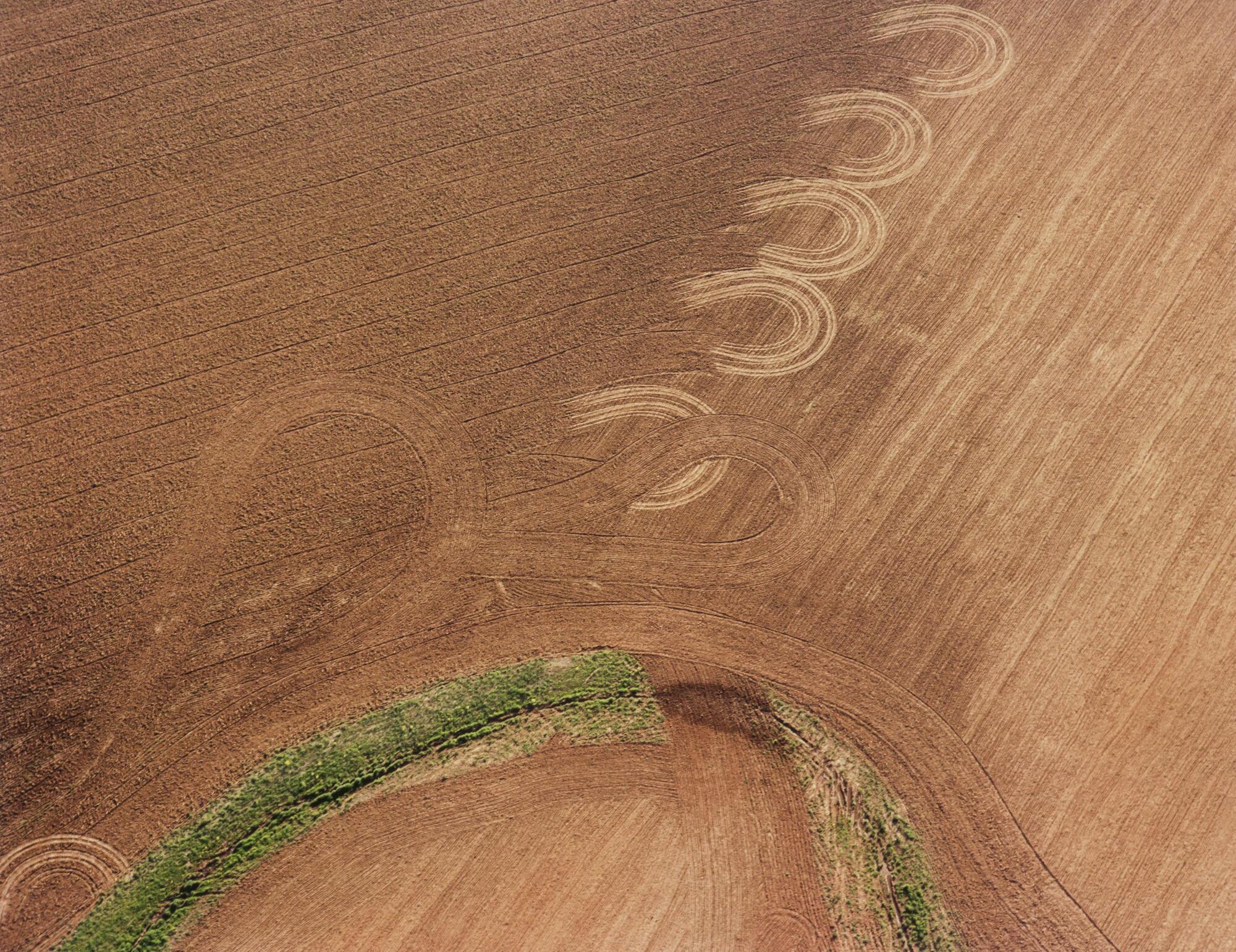 <br />
Print of an aerial view of plowed fields with U-shaped designs left by the machinery. A green swath cuts through the bottom fo the picture in another U-shape.
