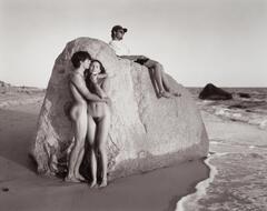 A photograph of a group of people on a beach, by a large rock. A nude couple leans against the rock, embracing one another. A clothed man sits on top of the rock looking out at the water. 