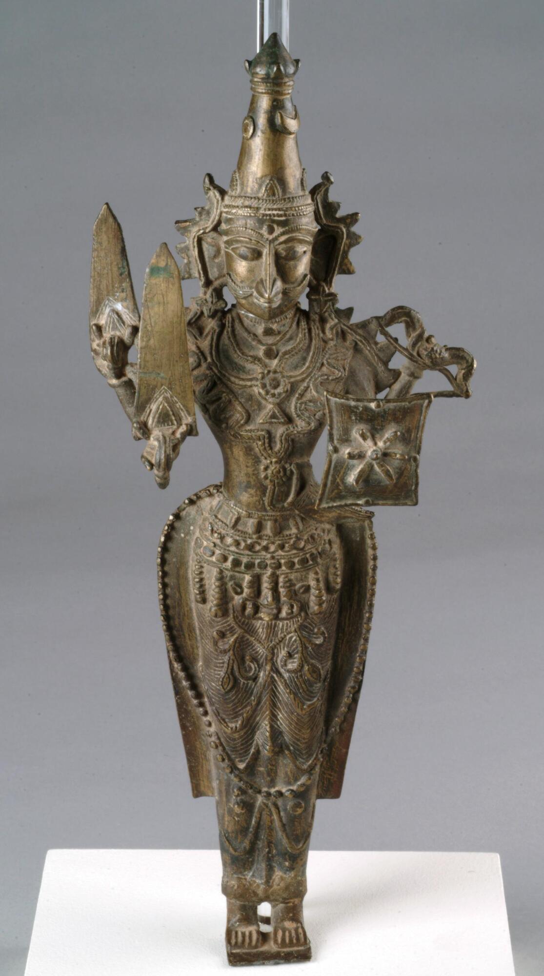 Shiva stands on a tiny base with little feet in a strict unbending stance.  The body is elongated with a small waist, the hips billowing out and tapering in a stylized way to the feet.  He has broad shoulders and has four arms.  Reading in a clockwise direction from the right front hand, he holds, a sword, another sword, a bow and a shield.  He wears a lot of jewelry, necklaces that cover his entire chest, at least three belts with pendant decorations and what appears to be a bustle like garment that billows out from his waist and has a beaded border that curves down across his legs below his knees.    He wears large earrings and a crown decorated with a disk for the sun and crescent for the moon.  He has large wide opened eyes and what almost looks like two sets of eyebrows, a dot where his third eye should be and a luxurious moustache.<br />