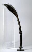 Wooden circular staff with a circular human face in the middle of the shaft. The bottom of the staff contains concentric rings that form a point at the base. The top of the staff has a black and white checkered pattern that wraps around the staff. Animal hairs stick out the top of the staff. 