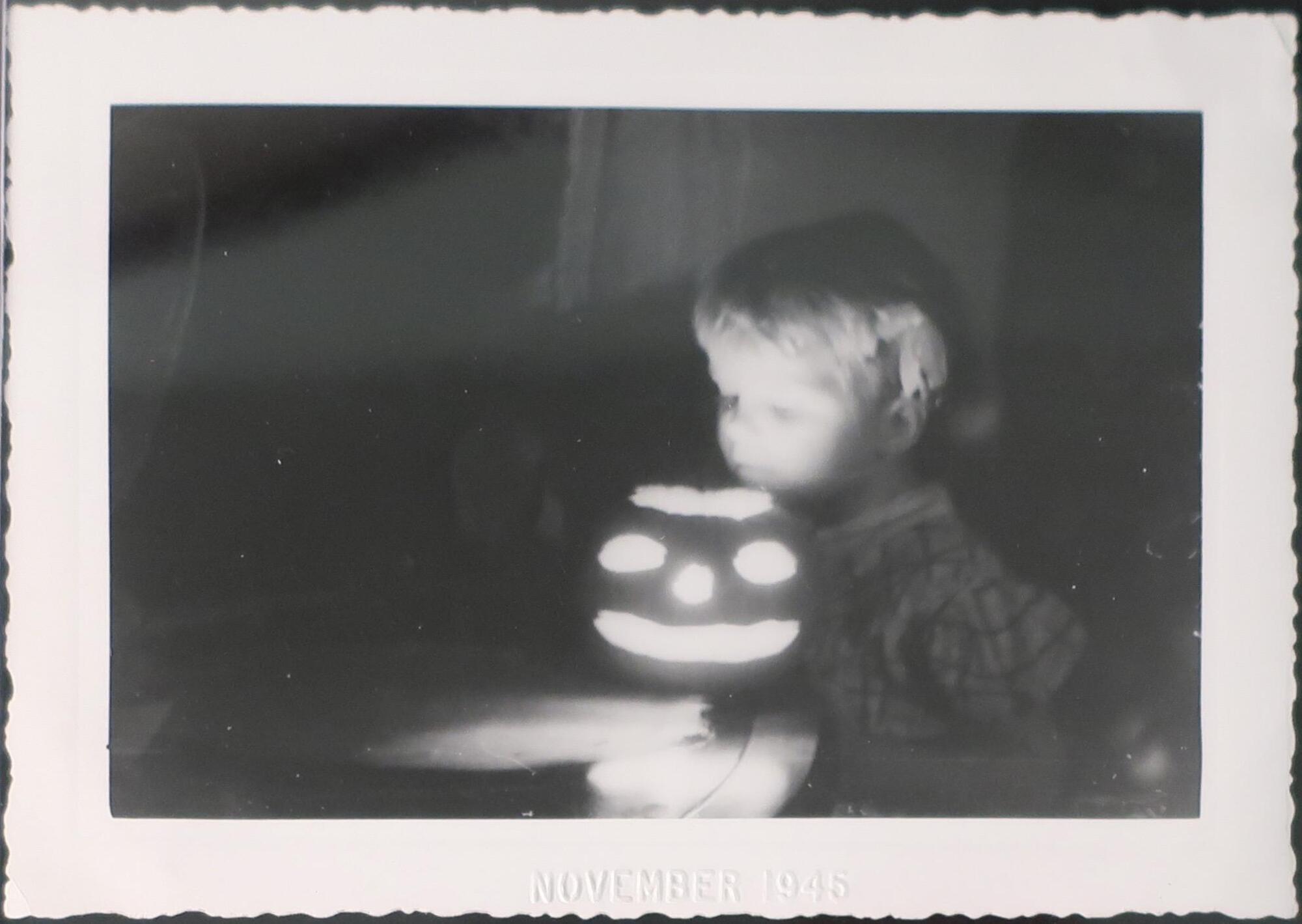 A young child peers down into the top of a lit jack-o&#39;-lantern, illuminating their face from below. Most of the image is in shadow, but the carved face of the pumpkin and the child&#39;s face are both brightly lit.