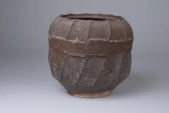 A container with a cylinder shaped body and a wide opening, it has a pattern of lines on the outer body. Dark brown coloring. Small series of lines near the top of the bowl that go all the way around. Rest of the bowl has slightly curved beveled edges that go from top ot bottom.
