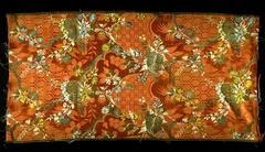 This brilliant coral-colored silk features an elaborate brocaded pattern consisting of exuberant sprays of flowers in yellow, blue, white, and gold thread joined to bunches of fruit and other ornament woven with silver thread. The ground is enlivened by a damask pattern of more schematic vegetal and geometric ornament that echoes these dominant motifs.